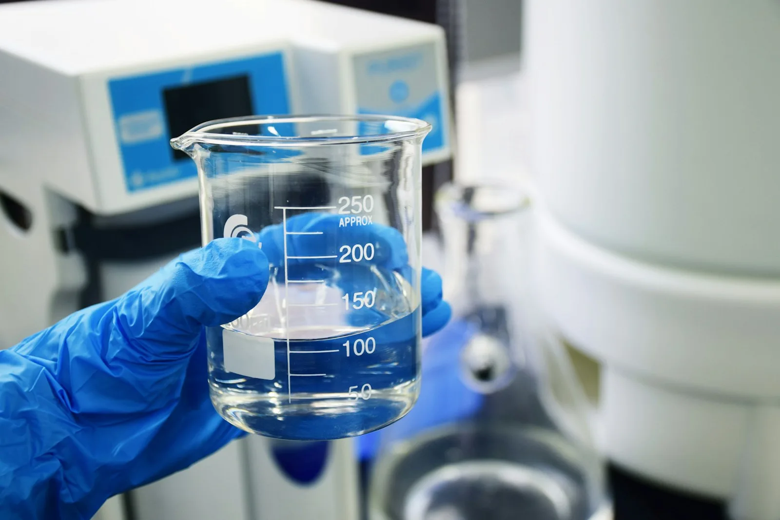 Mano con guante azul sosteniendo un vaso de precipitados con agua transparente en un laboratorio de análisis